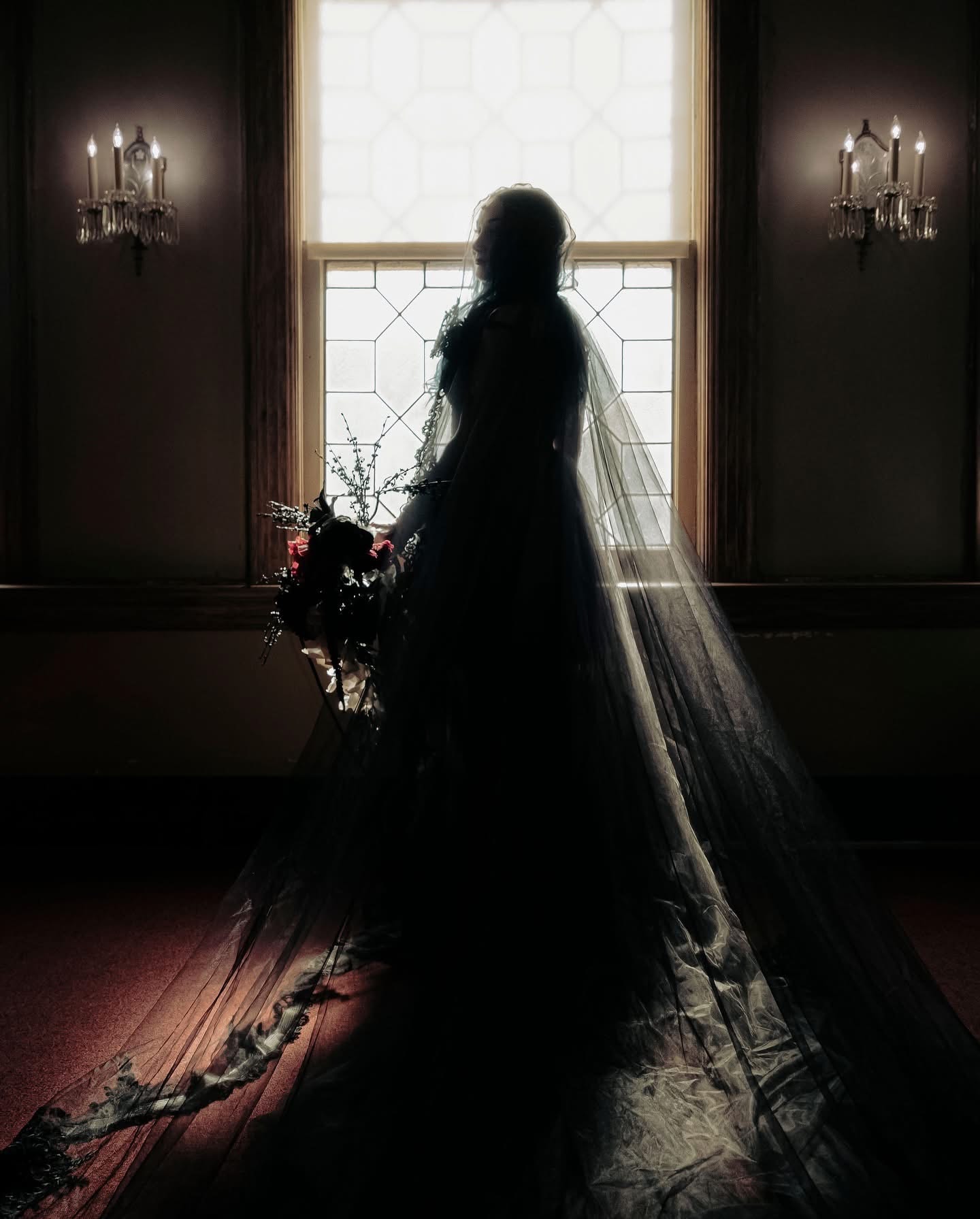 Bride silhouetted at window in black gown with dark floral bouquet